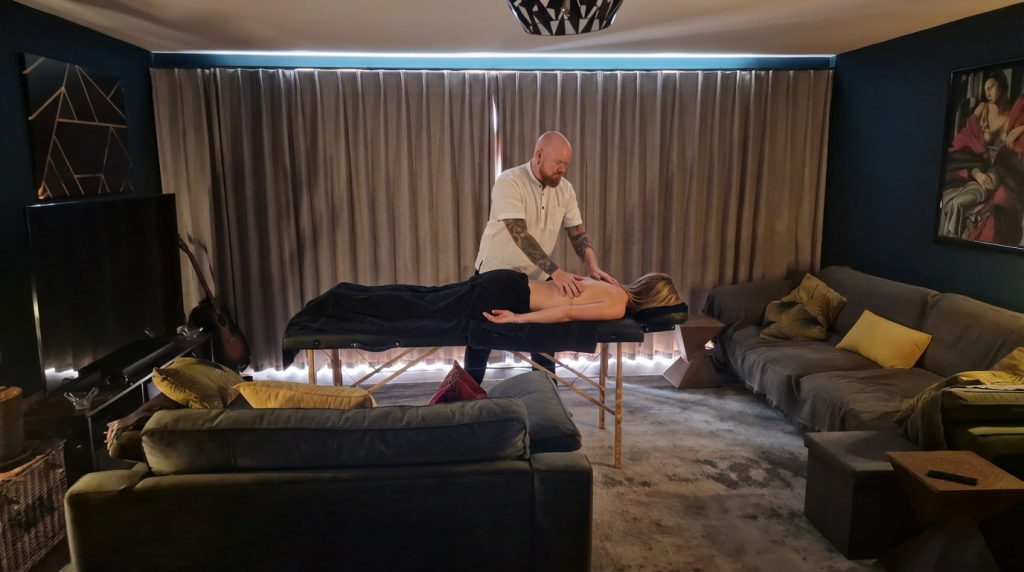 Masseur giving back massage to a person lying on a massage table in a cozy living room with couches in view.